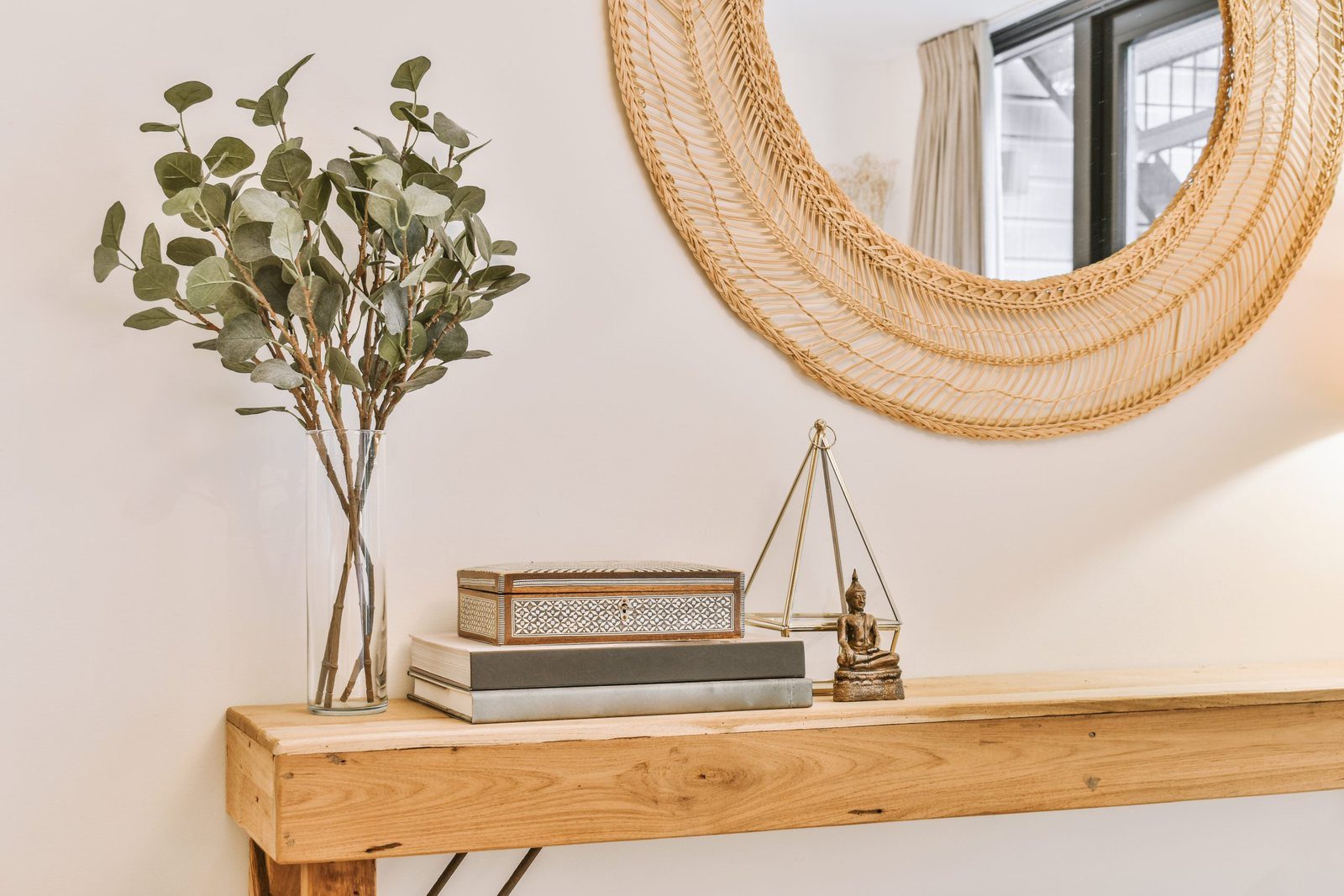 A wooden console table with eucalyptus in a glass vase, stacked books and a round rattan mirror above.